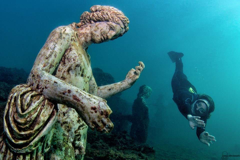 Underwater statue of a seated figure reaching toward a scuba diver swimming nearby