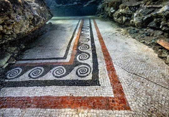 Ancient mosaic walkway with black-and-white spiral border and red lines between rocky walls