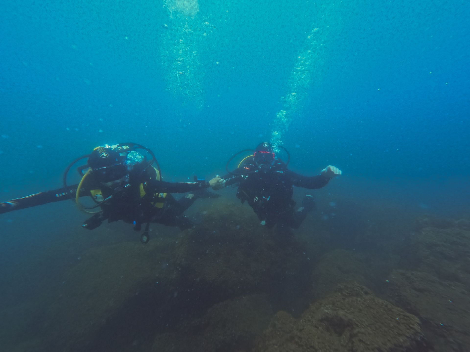 Two scuba divers swim underwater over a rocky seabed in blue water.