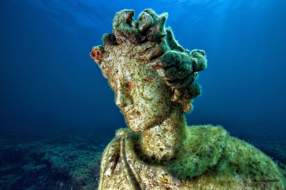 Underwater statue covered in algae, with blue ocean background