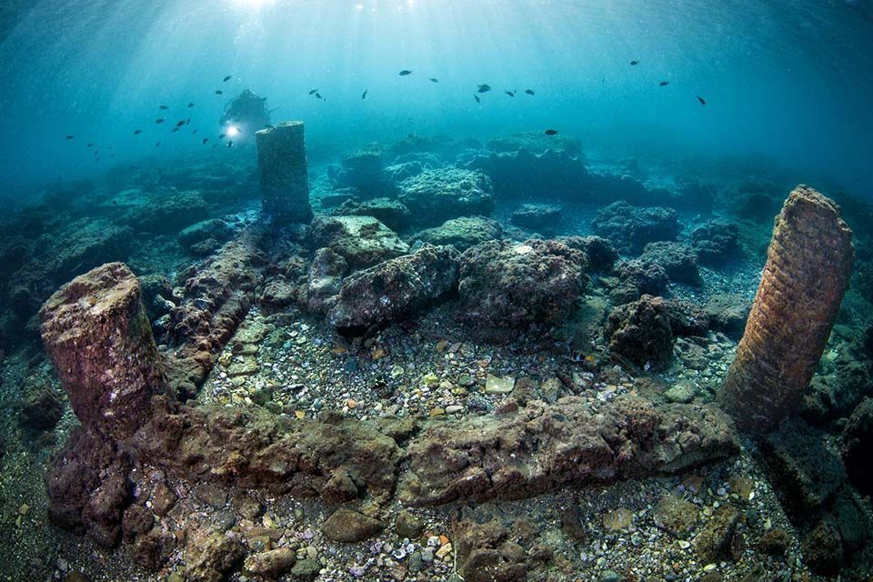 Underwater view of a sunken stone ruin with coral-covered pillars and blue light filtering through the water