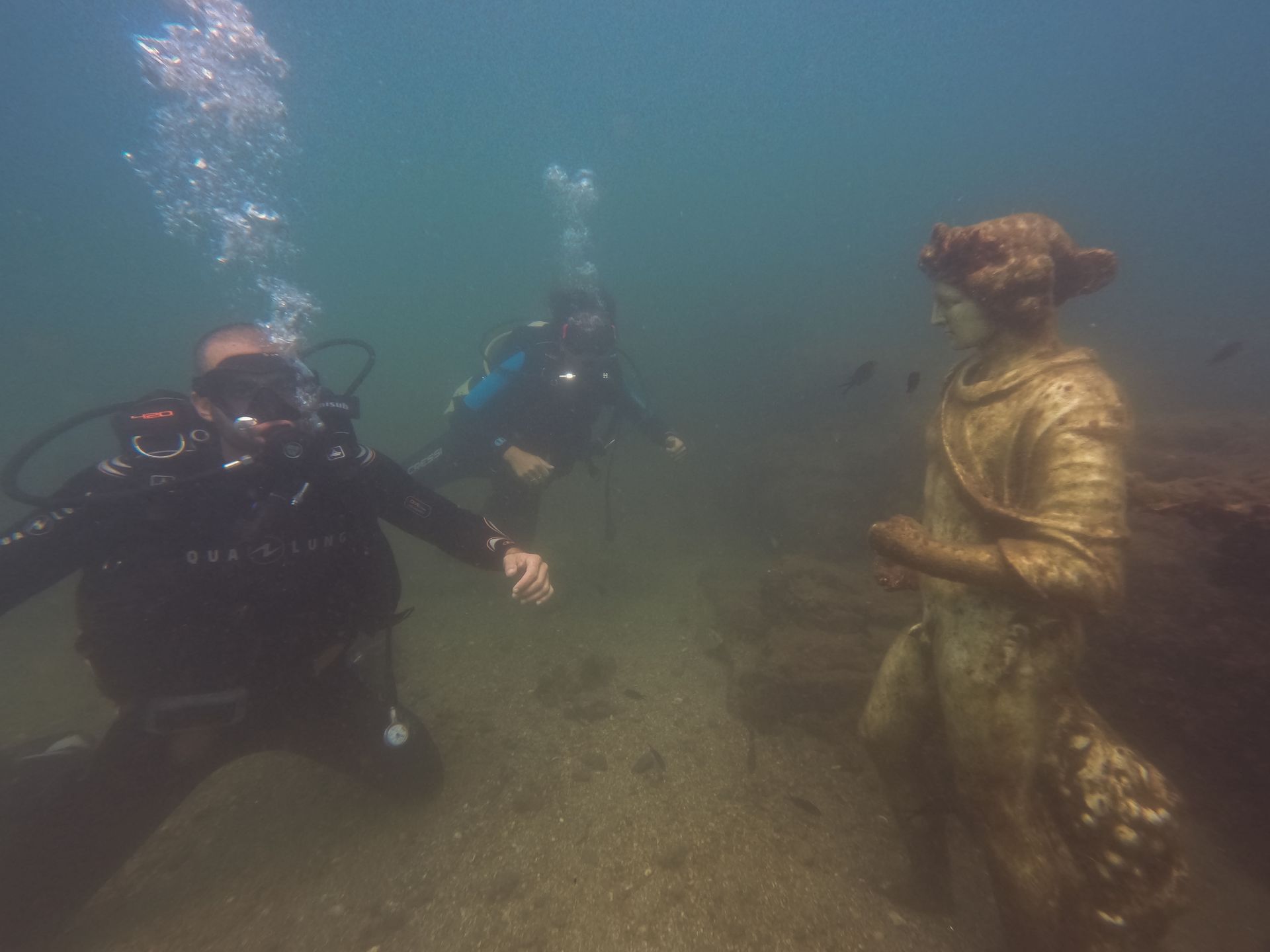 Divers underwater, one in a gold suit, facing a statue in a murky seafloor setting