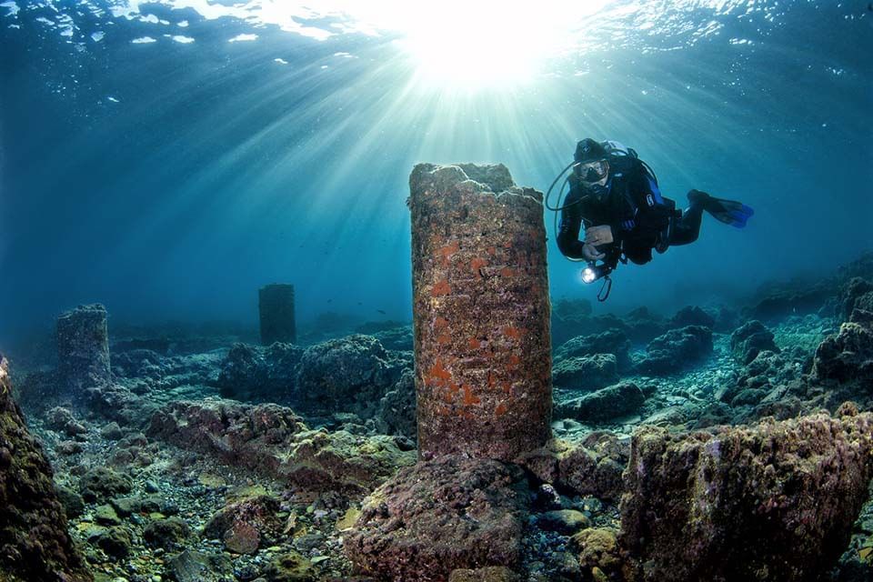 Scuba diver exploring an underwater ruins site with sun rays and coral-covered stone pillars