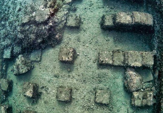 Aerial view of a shallow green-blue ruin with scattered stone blocks and walls underwater