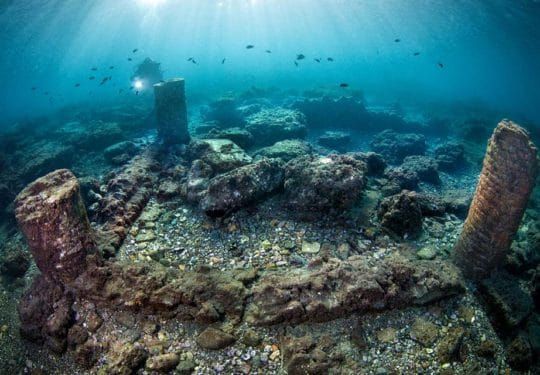 Underwater ruins of a stone structure on a rocky seafloor with blue sunlight filtering through water