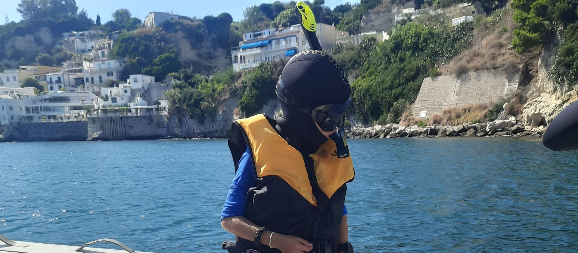 Person in a yellow life vest on a boat with blue water and a rocky shoreline in the background