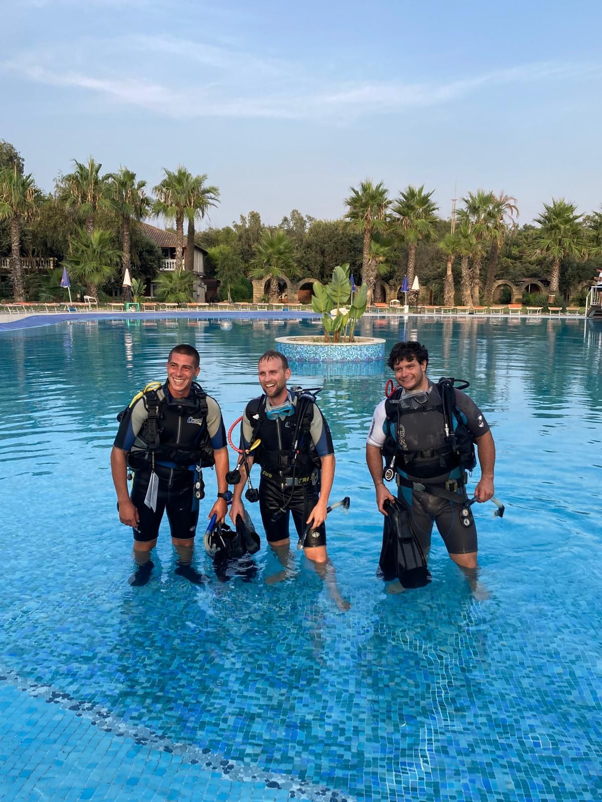 Three scuba divers standing in a blue resort pool with palm trees in the background
