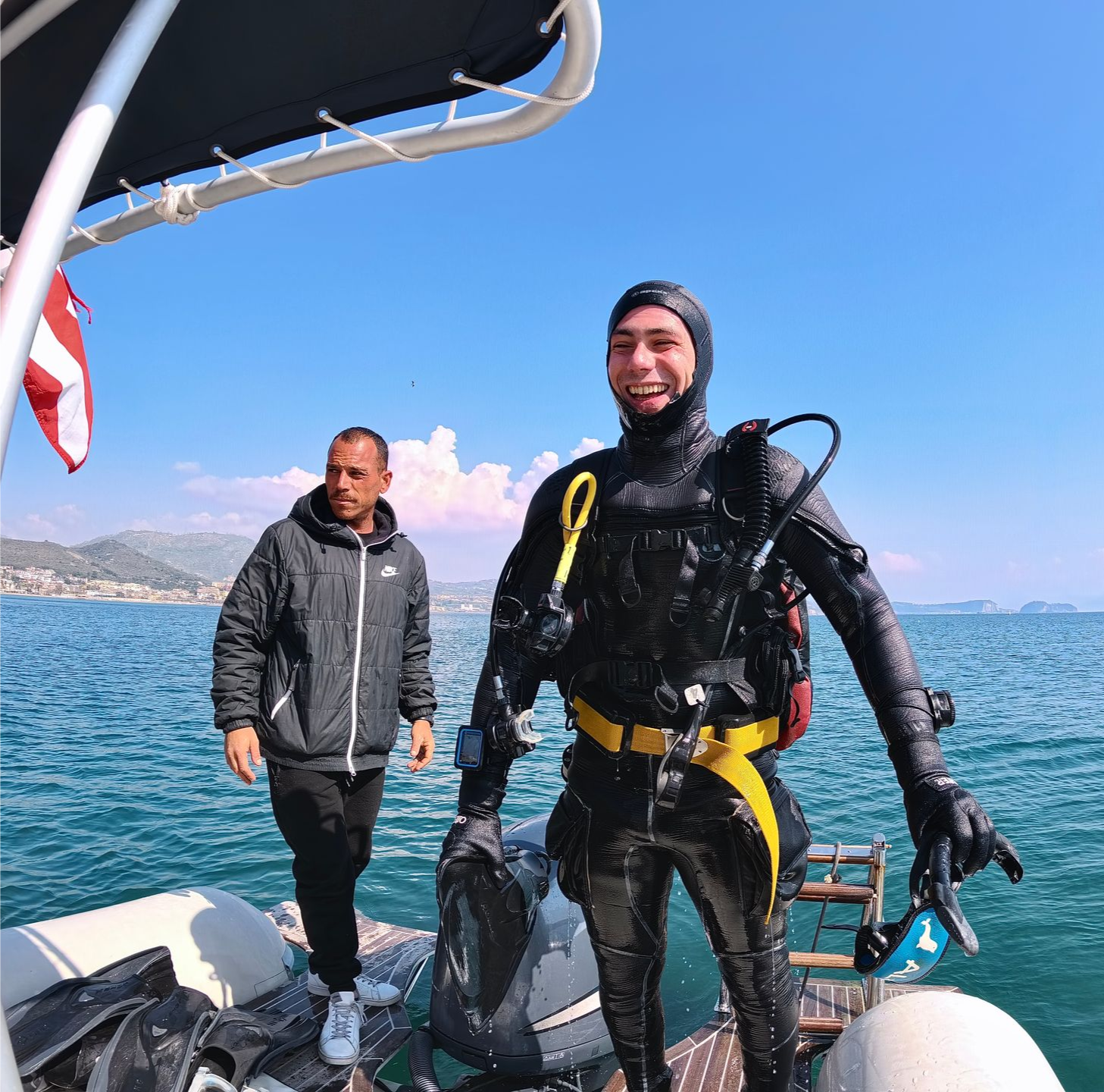 Two people on a boat deck by the sea, one in a wetsuit smiling, with a clear blue sky.