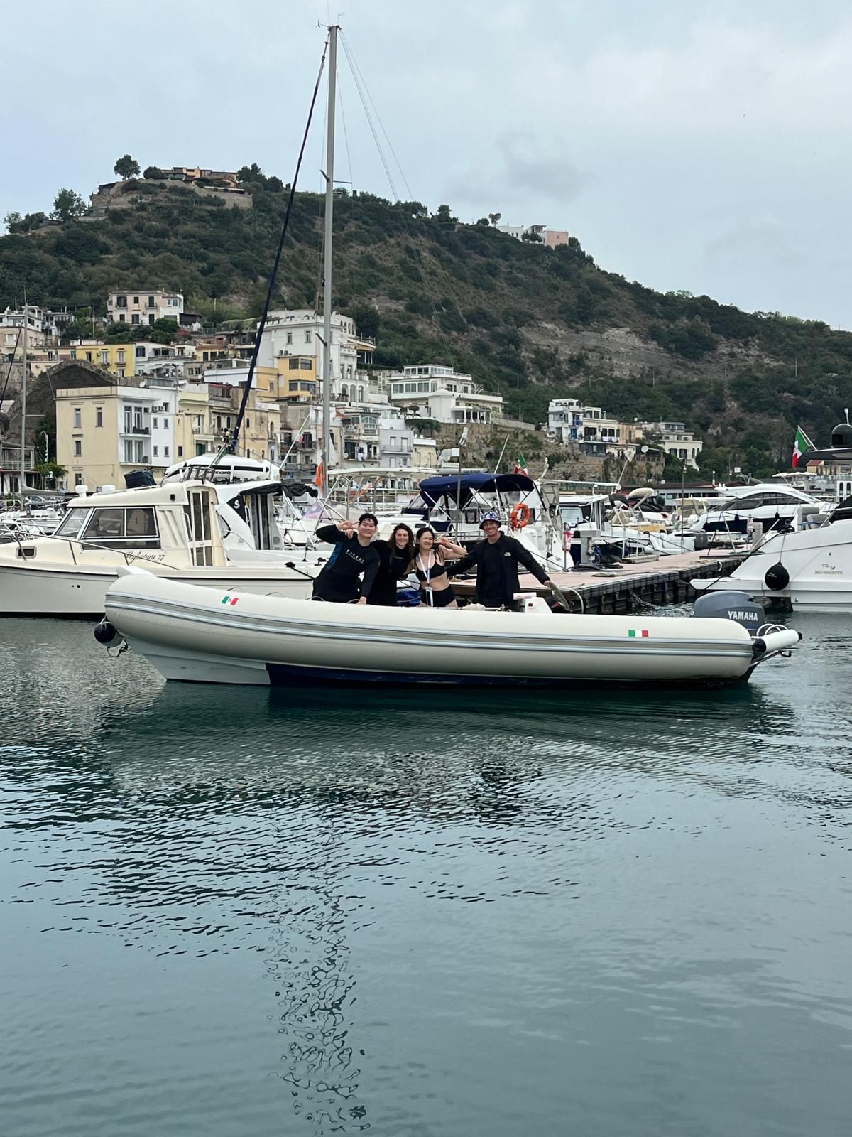 Small motorboat with people on calm harbor water, with yachts, hillside buildings, and a cloudy sky behind it