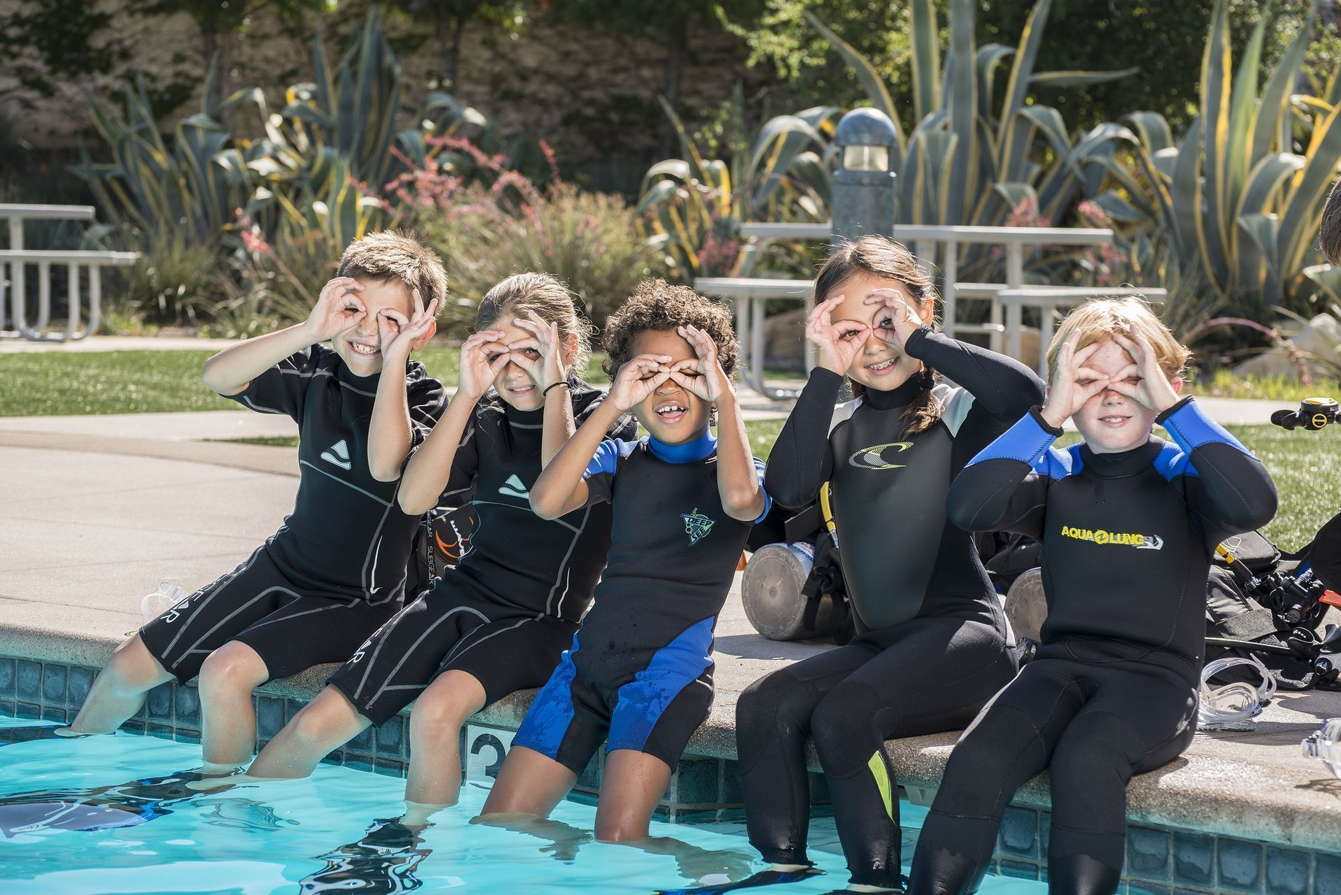 Five children in wetsuits smiling and making goggles with their hands while sitting by a pool