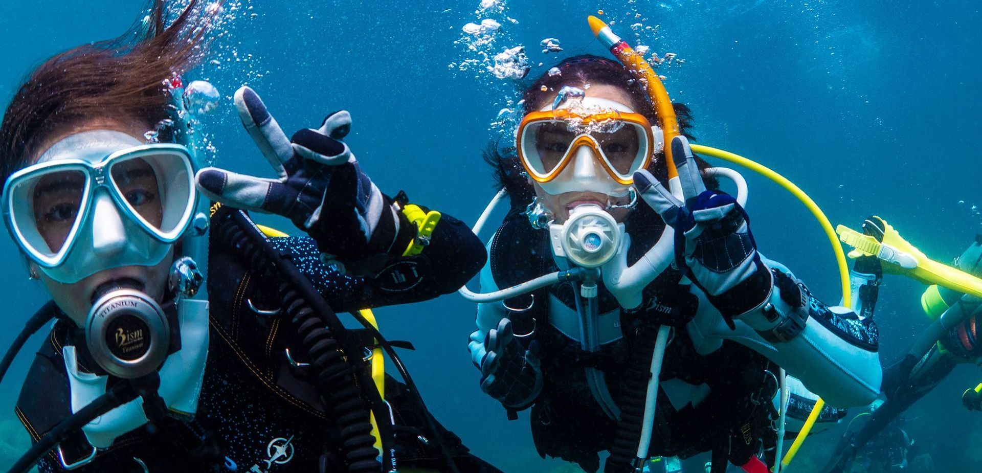 Two scuba divers underwater giving thumbs-up, wearing masks and bright gear, surrounded by blue water and fish