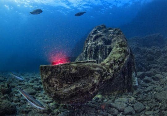 Submerged stone sculpture on a rocky seafloor, with a red glow and fish swimming above
