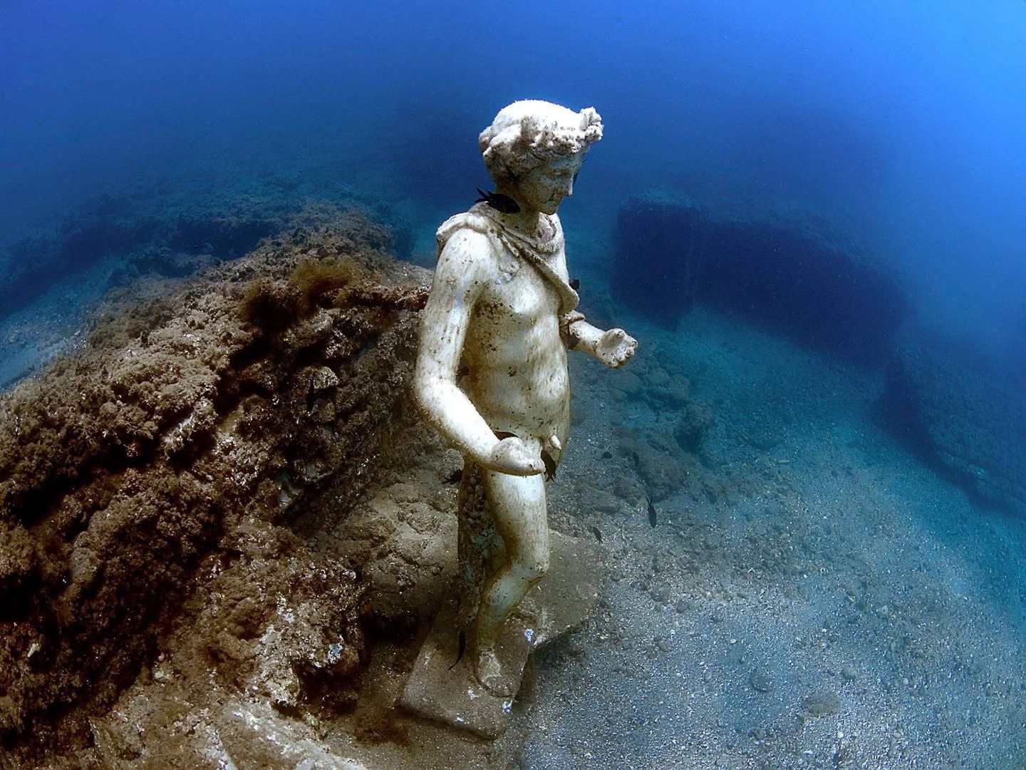 Submerged stone statue underwater beside a coral-covered reef in blue ocean water