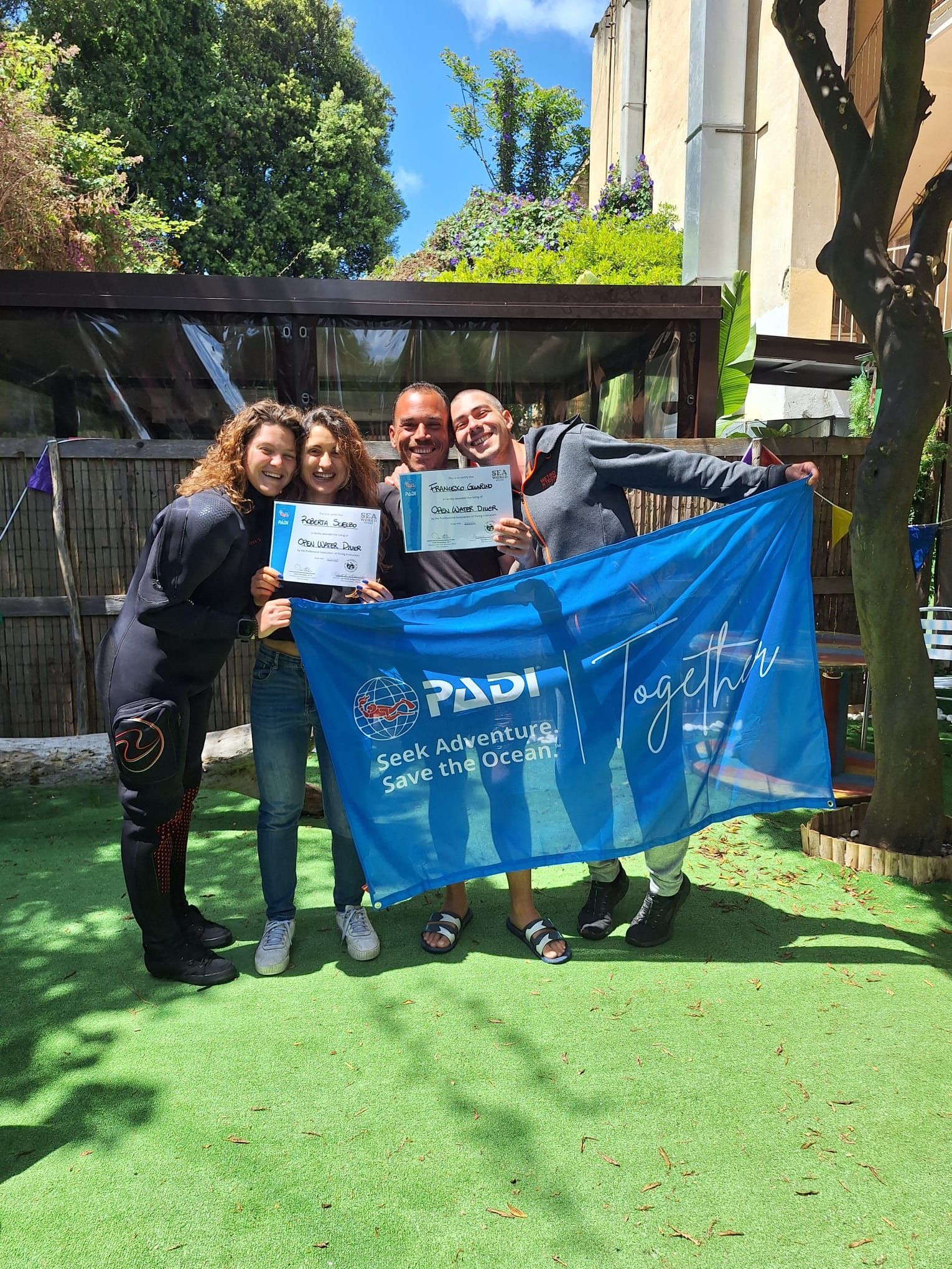 Four people posing outdoors holding certificates and a blue banner on a sunny lawn