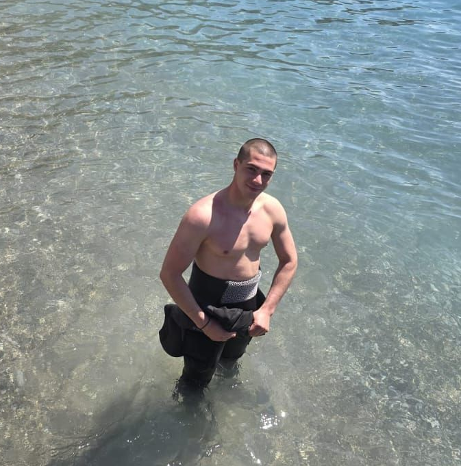 Man standing in shallow clear water, shirtless, wearing dark shorts, looking at the camera.