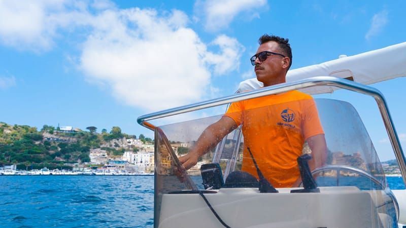 Man steering a boat on blue water with a coastal town in the background