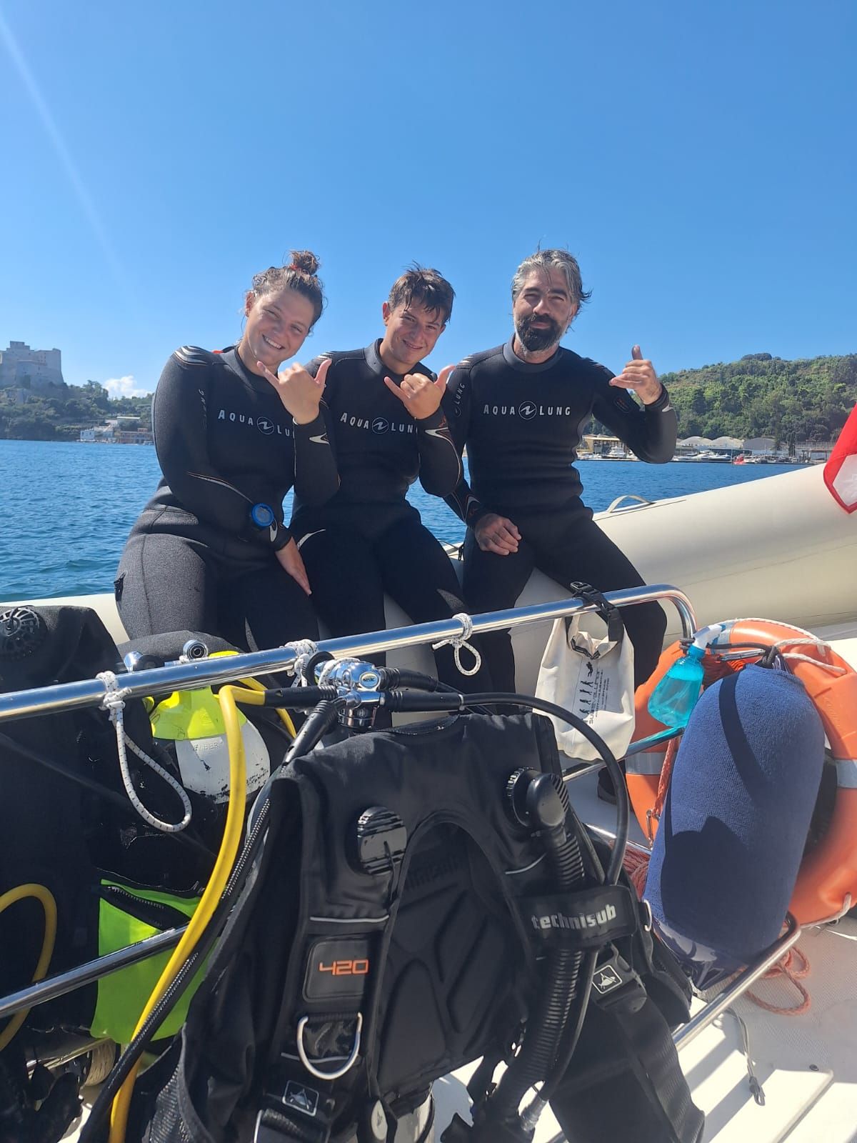 Three people in wetsuits smiling on a boat in bright sun, with blue water and shoreline behind them