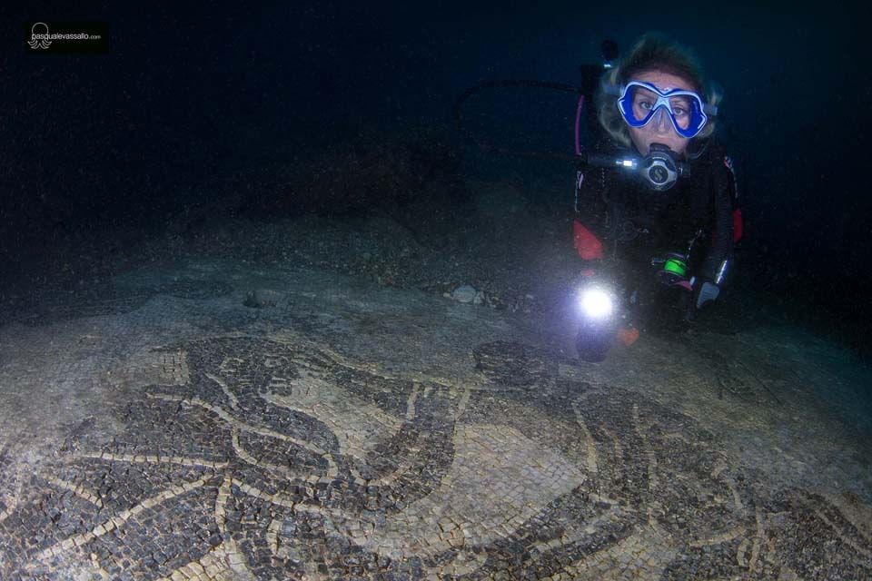 Scuba diver shining a flashlight over a dark seafloor with a large etched design.