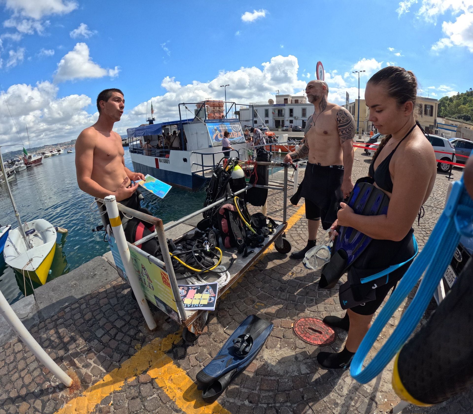 People in scuba gear preparing on a dock beside a harbor under a blue sky