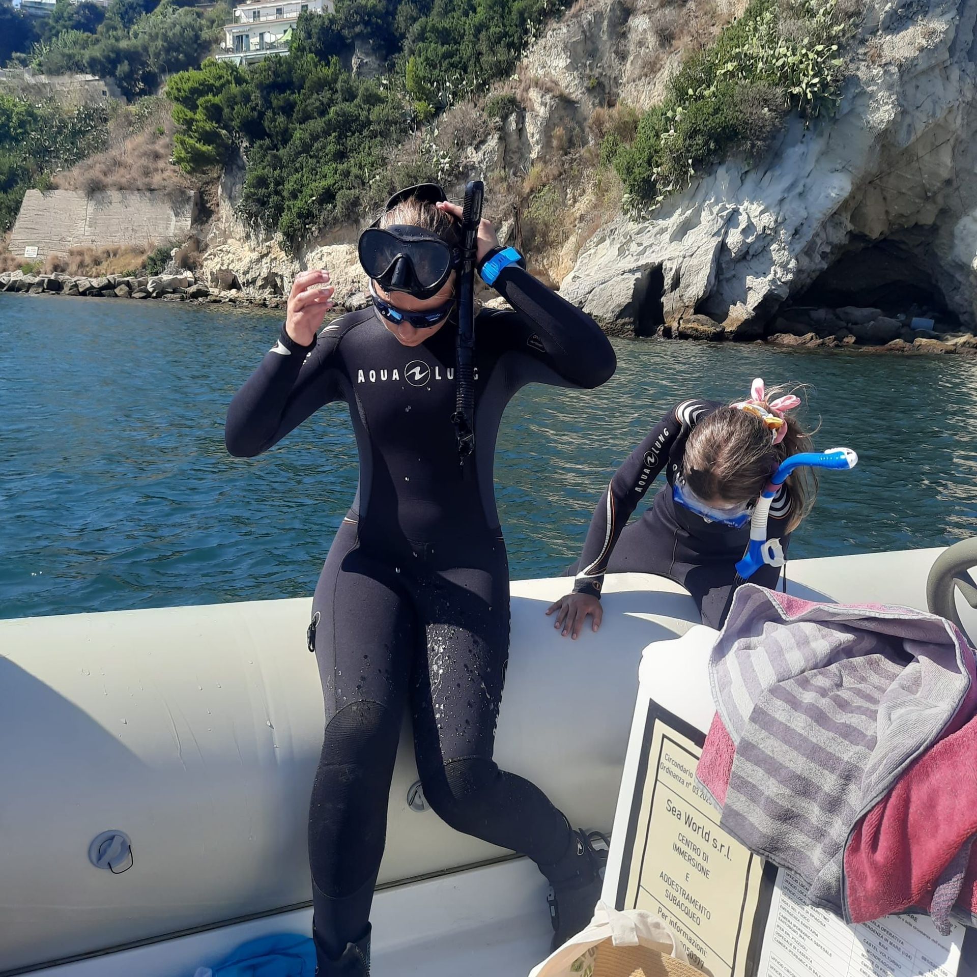 Two snorkelers on a boat, adjusting masks near rocky cliffs and blue water.