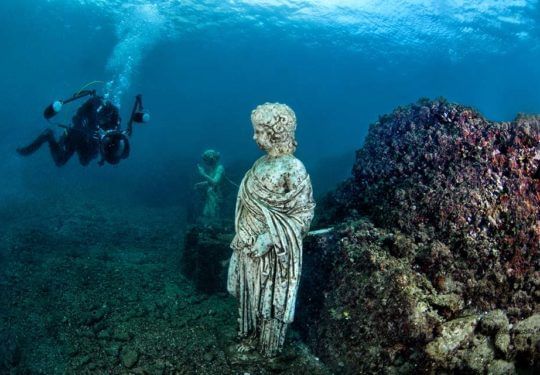 Diver exploring an underwater statue beside a coral-covered reef in blue water