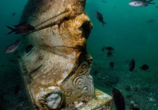 Underwater close-up of a weathered stone statue with fish swimming in teal water