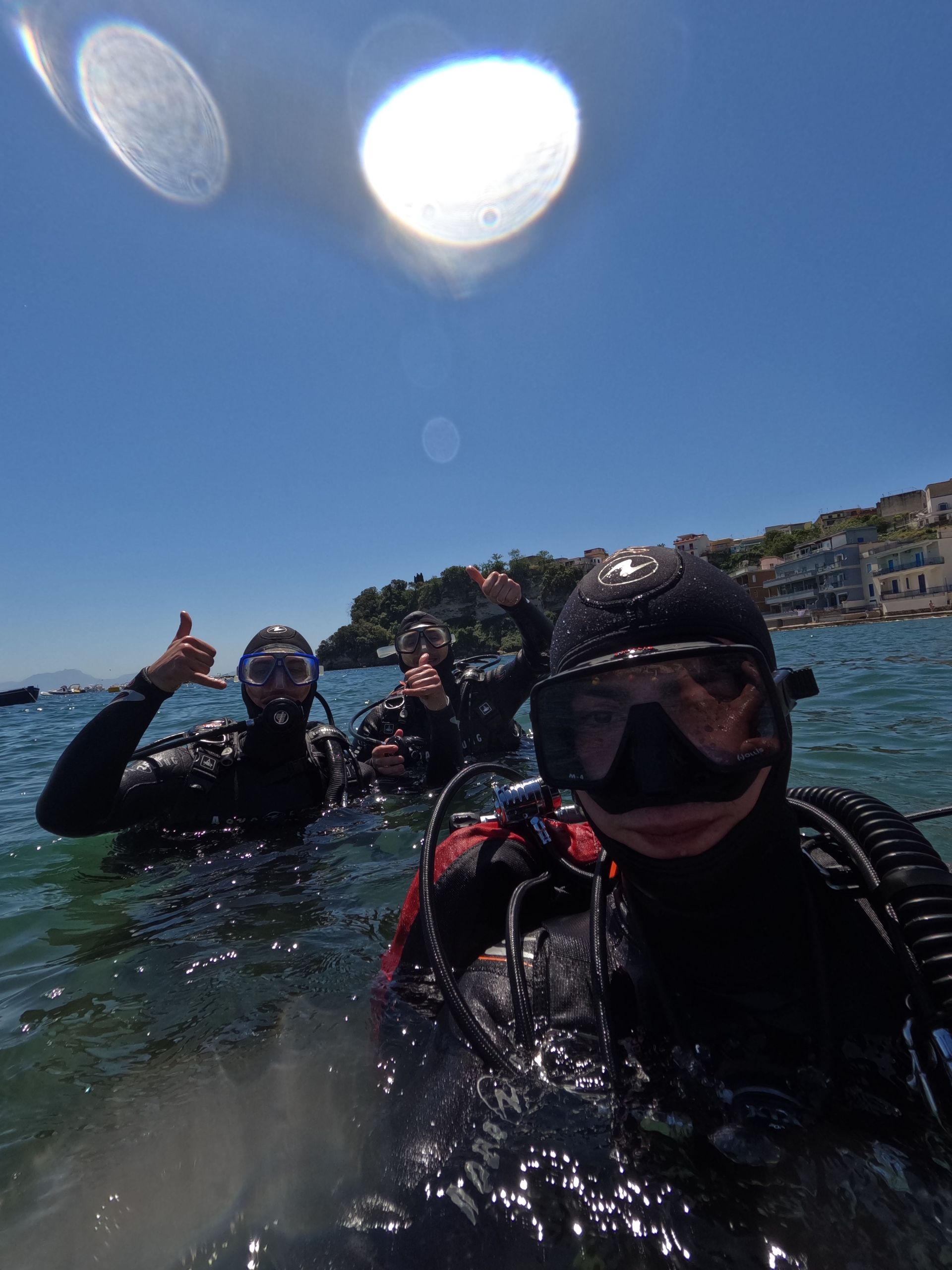 Three divers in the ocean near a rocky shoreline, posing with thumbs up under bright sun.