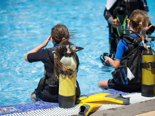 Two scuba divers in wetsuits sit by a pool with tanks and gear, preparing for a dive.