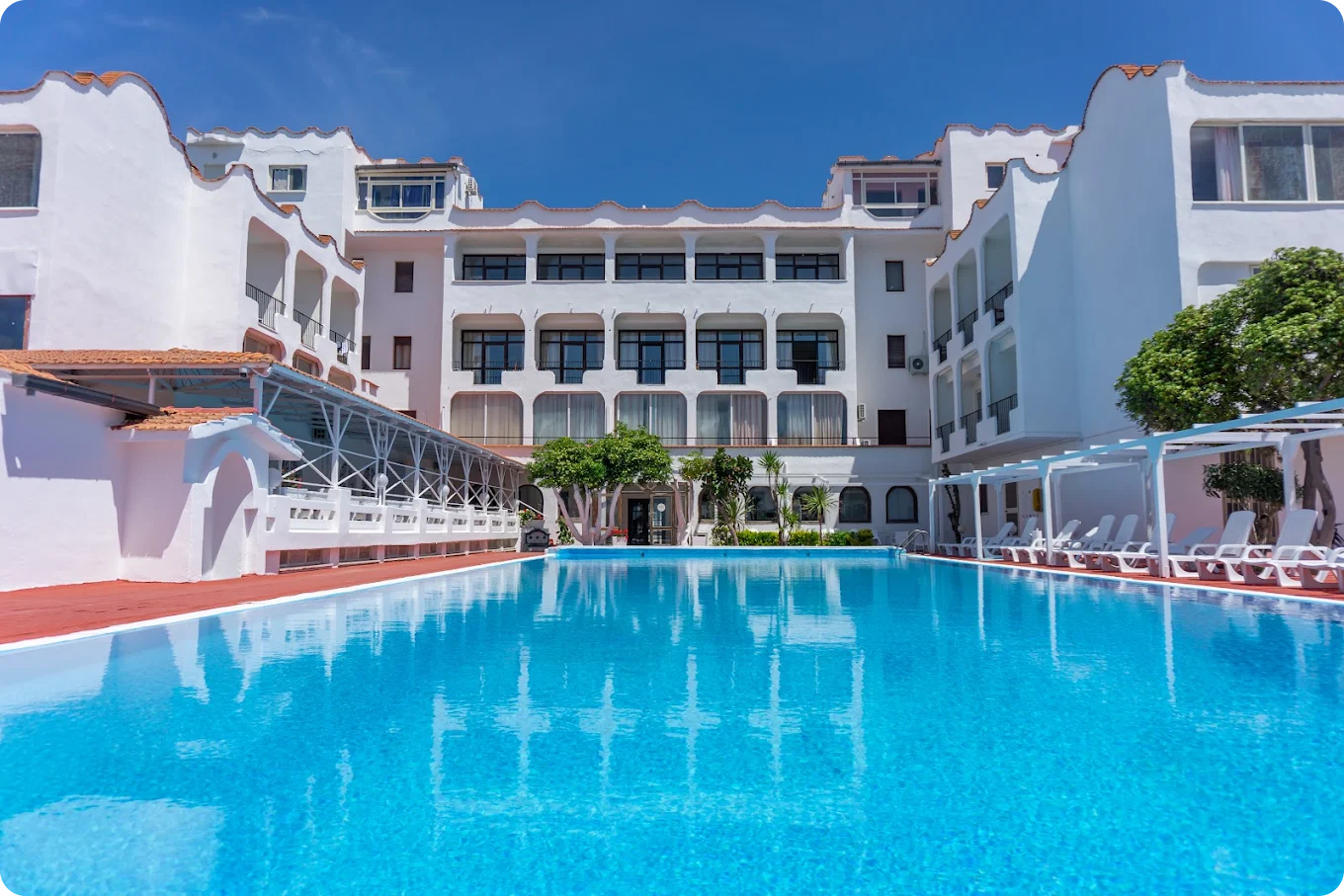 Large white hotel with balconies overlooking a bright blue swimming pool on a sunny day