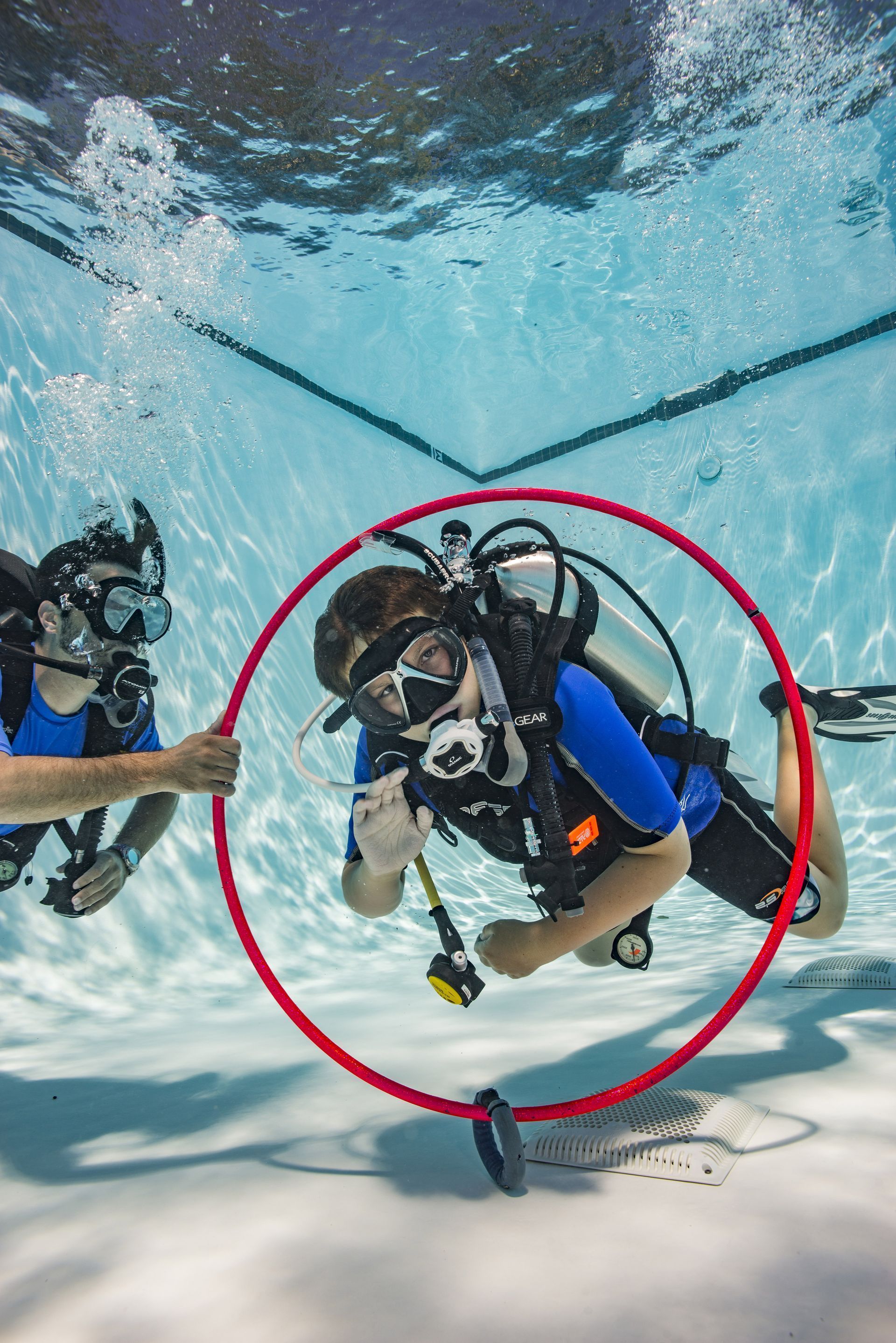 Scuba diver underwater in a red circle, holding a camera near the sandy pool bottom