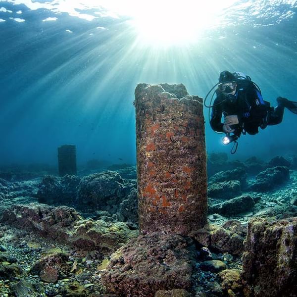 Underwater diver exploring a sunlit shipwreck beside a coral-covered pillar