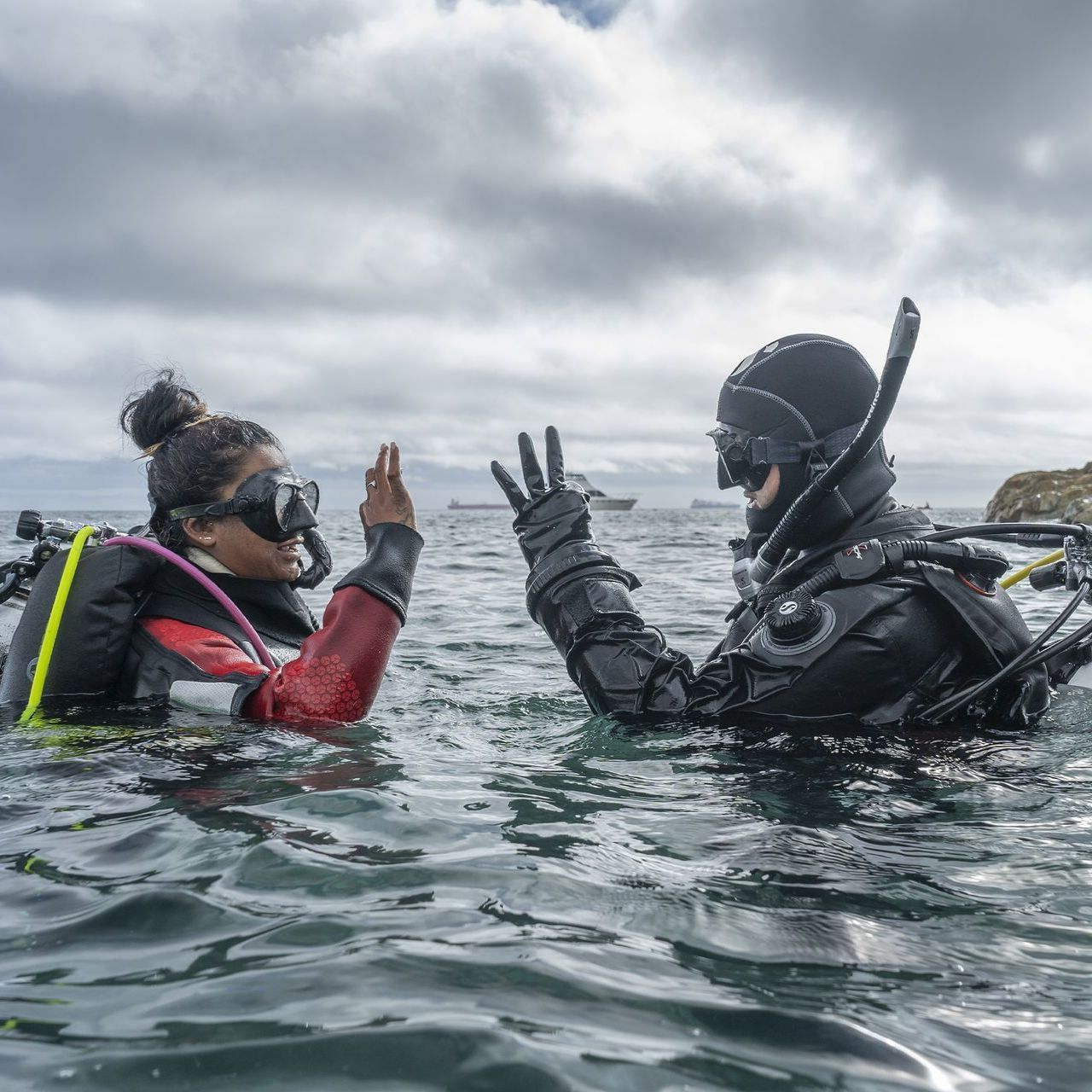 Two scuba divers in wetsuits underwater at the surface, facing each other with raised hands in a gray sea.