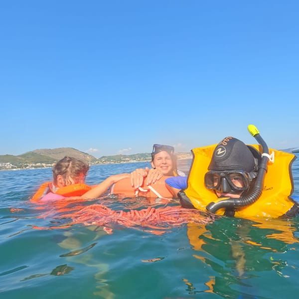 Three snorkelers float in blue water, one in a yellow vest and mask, with a sunny coastline behind them.