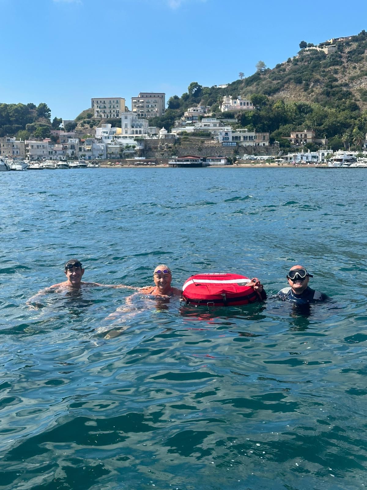 Three swimmers in open water with a red float ring near a rocky shoreline and hillside buildings