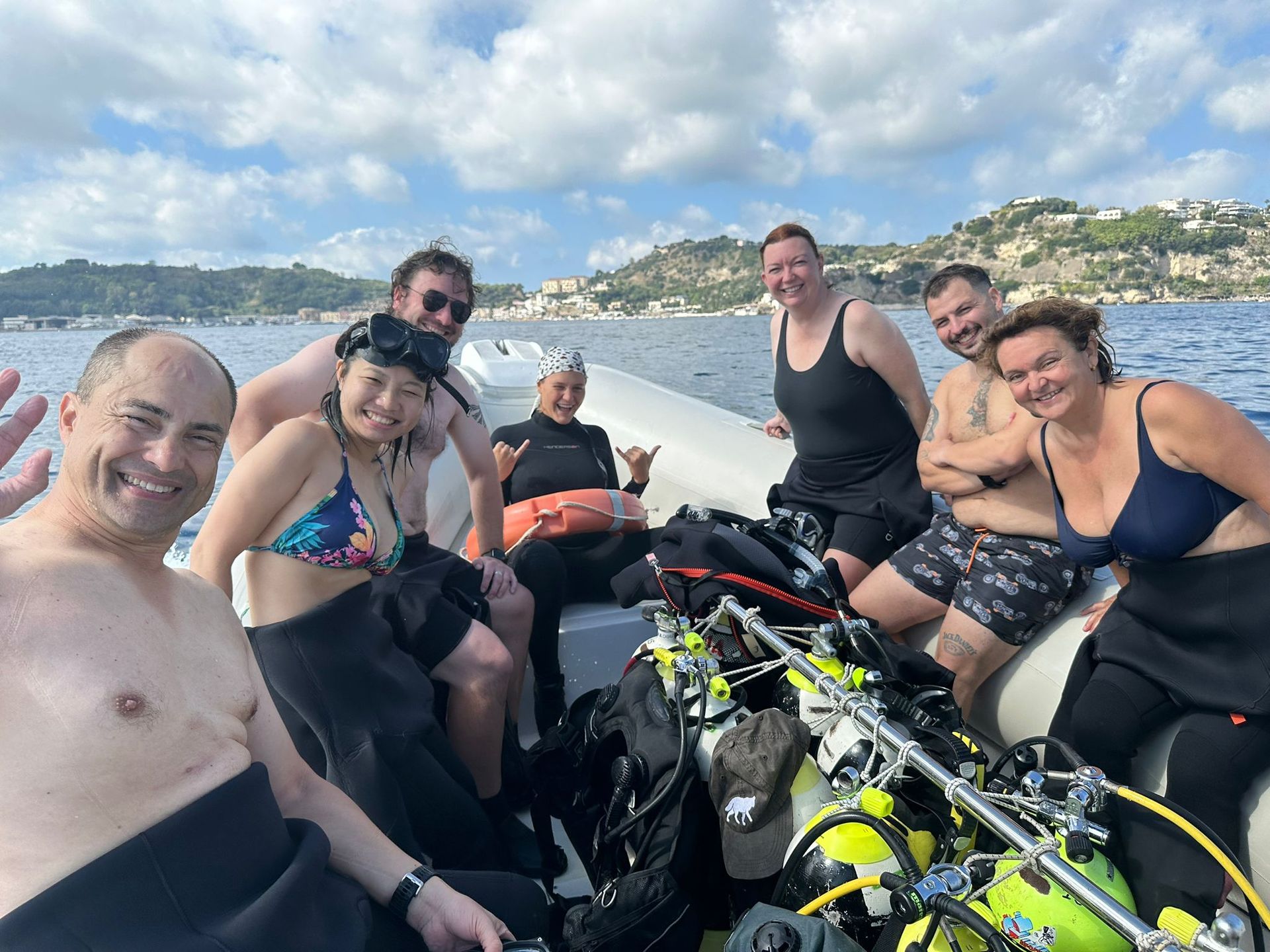 Group of friends on a boat with scuba gear, smiling on the water near a coastal hillside