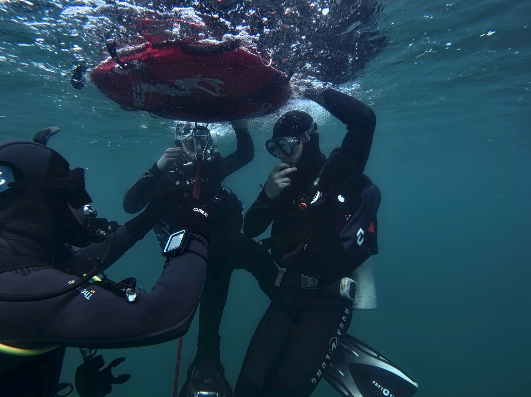 Scuba divers underwater releasing a red bag, with one diver signaling and another filming nearby