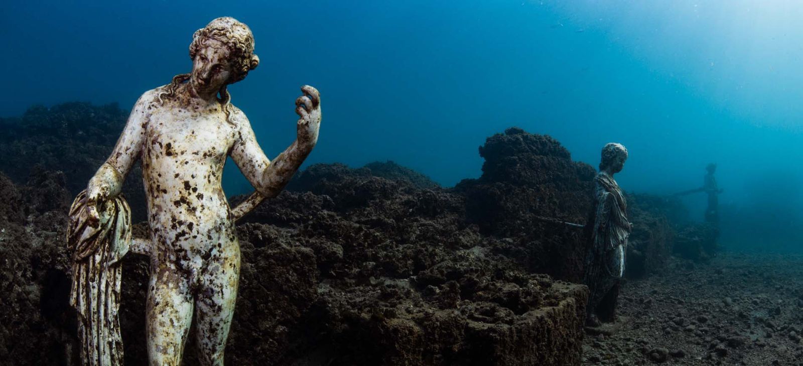 Underwater statues on a rocky seabed, one in the foreground raising a hand, with blue water and a distant figure