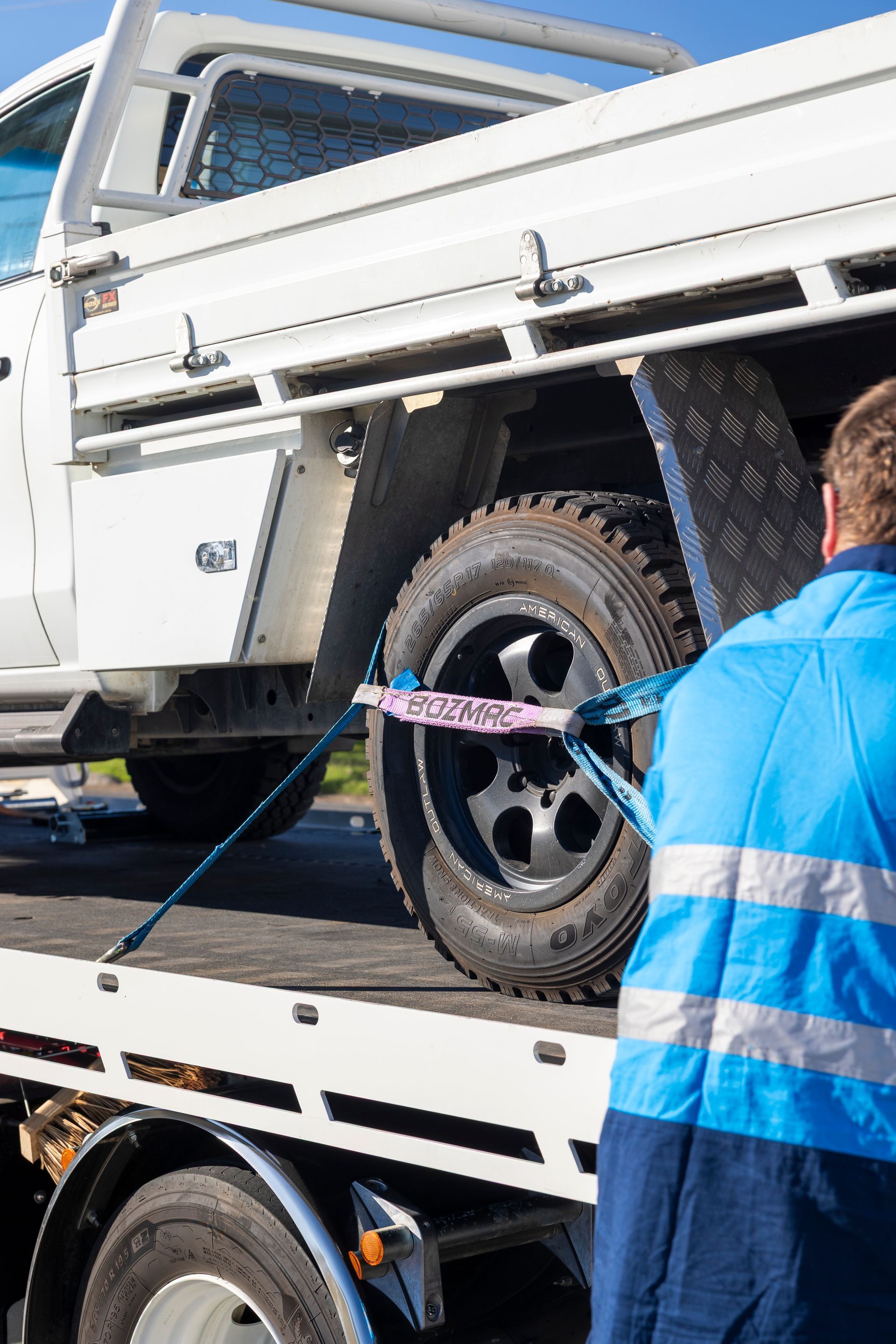 Person securing a white truck's tire to a tow truck with a blue strap, outdoors, sunny.— Perlyn Towing  in Gladstone, QLD