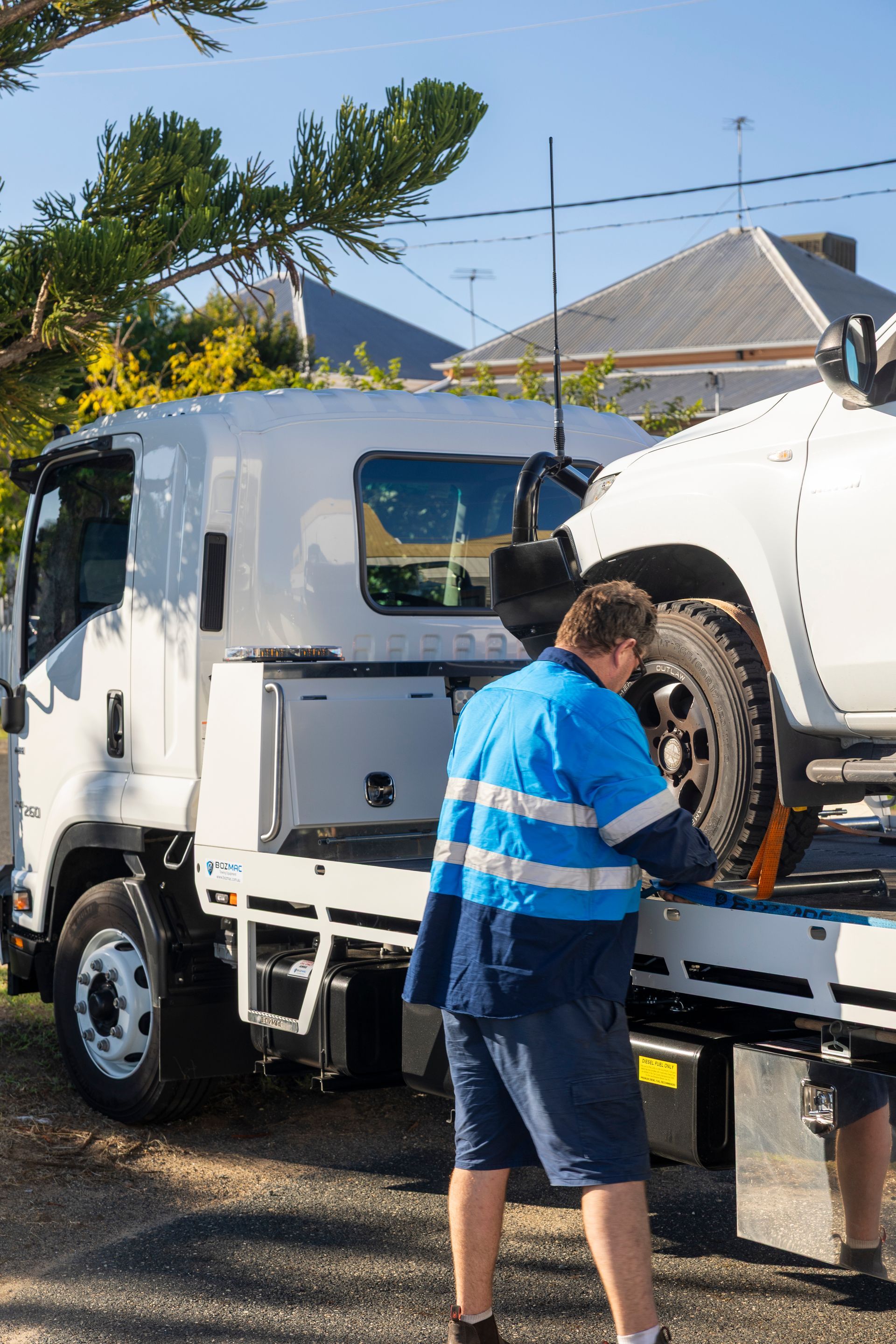 A tow truck operator in blue and white work clothes secures a white vehicle on a tow truck in front of a house.— Perlyn Towing  in Rockhampton City, QLD