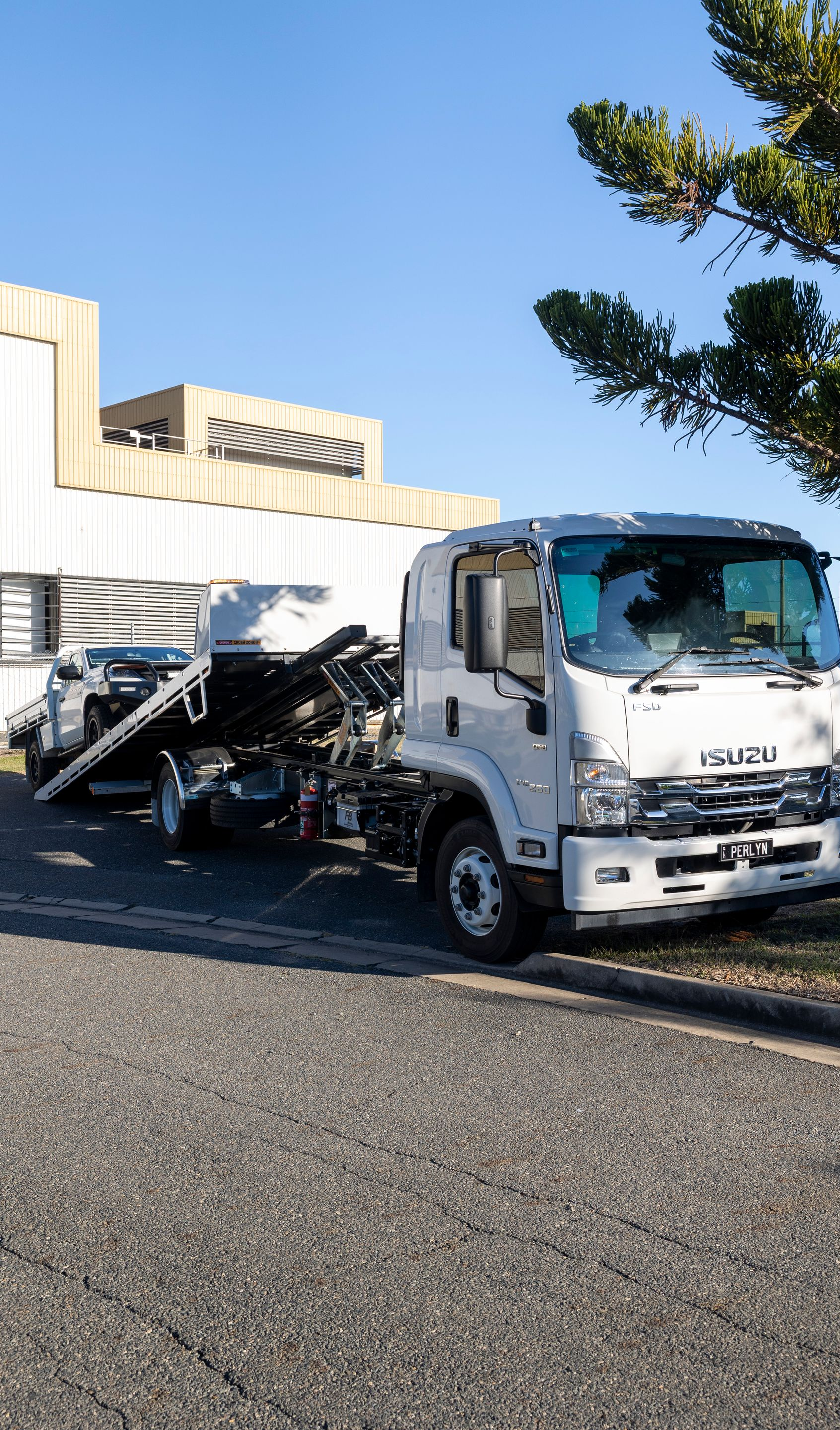 White Isuzu tow truck parked on asphalt in front of a building, with a damaged vehicle on the flatbed.— Perlyn Towing in Rockhampton City, QLD