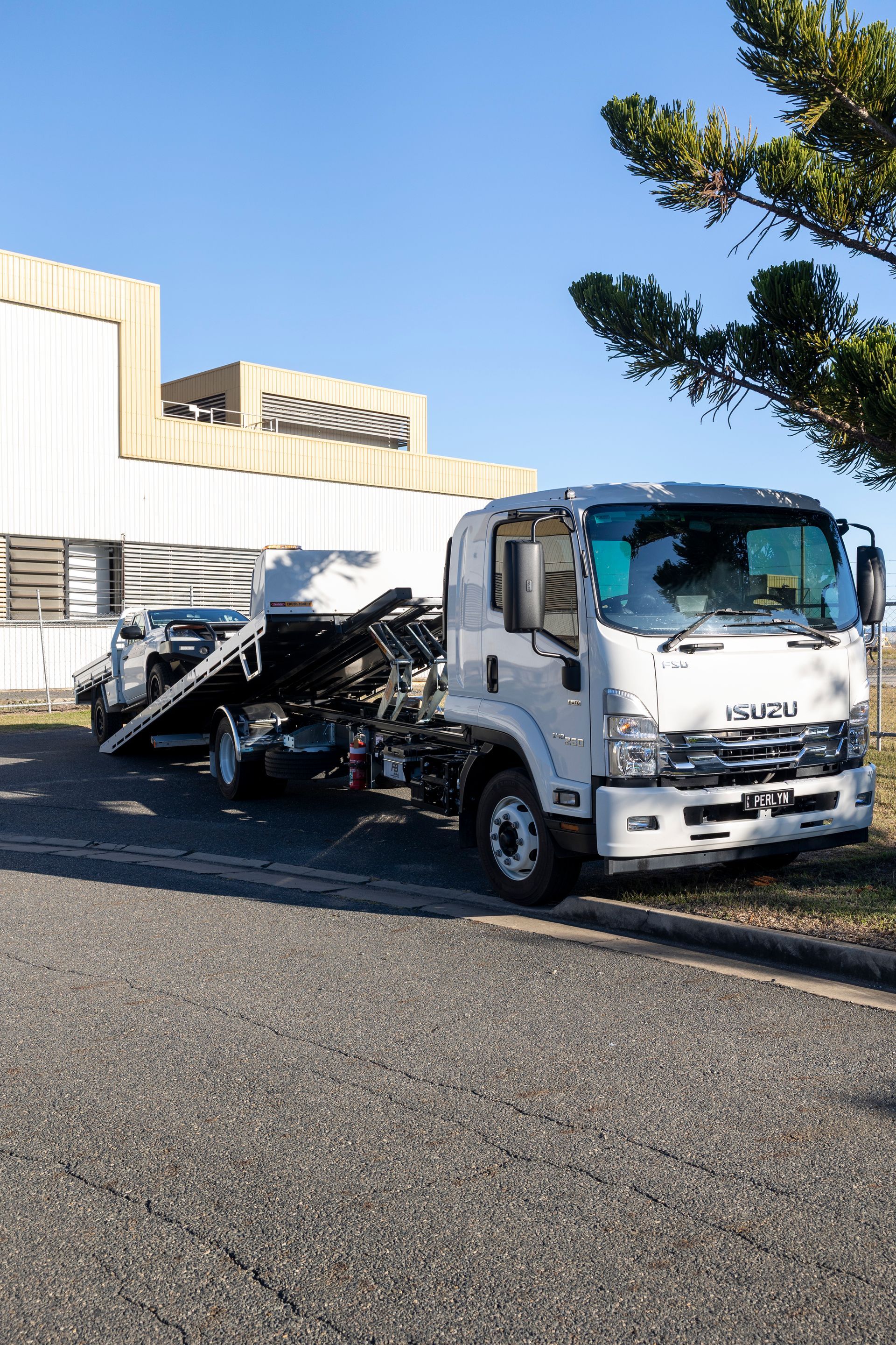 White Isuzu tow truck with a damaged car on its flatbed; parked on a paved area beside a building.— Perlyn Towing in Emerald, QLD