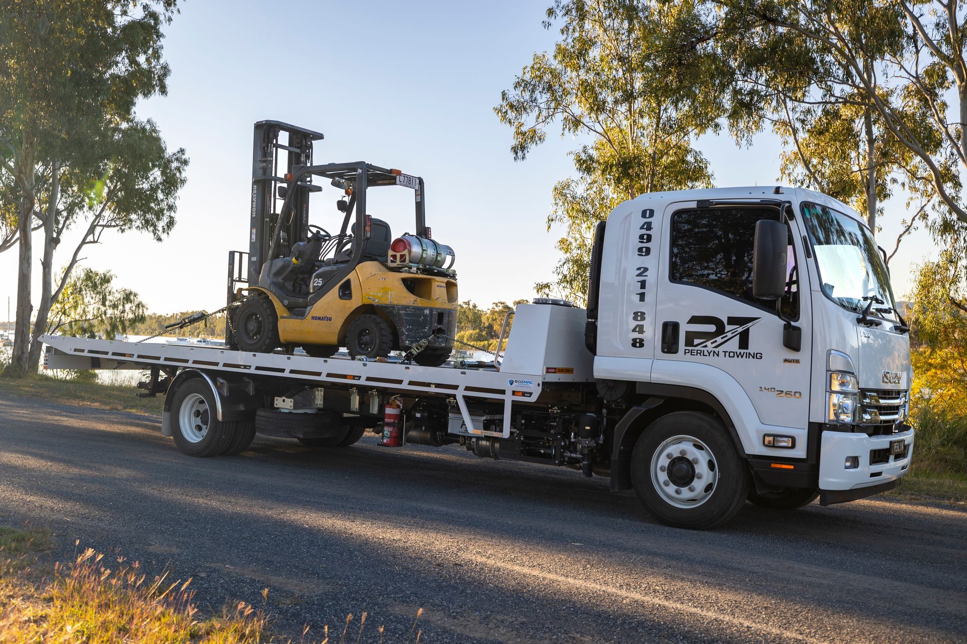 White flatbed truck transporting a yellow forklift on a road by trees and water.— Perlyn Towing in Emerald, QLD
