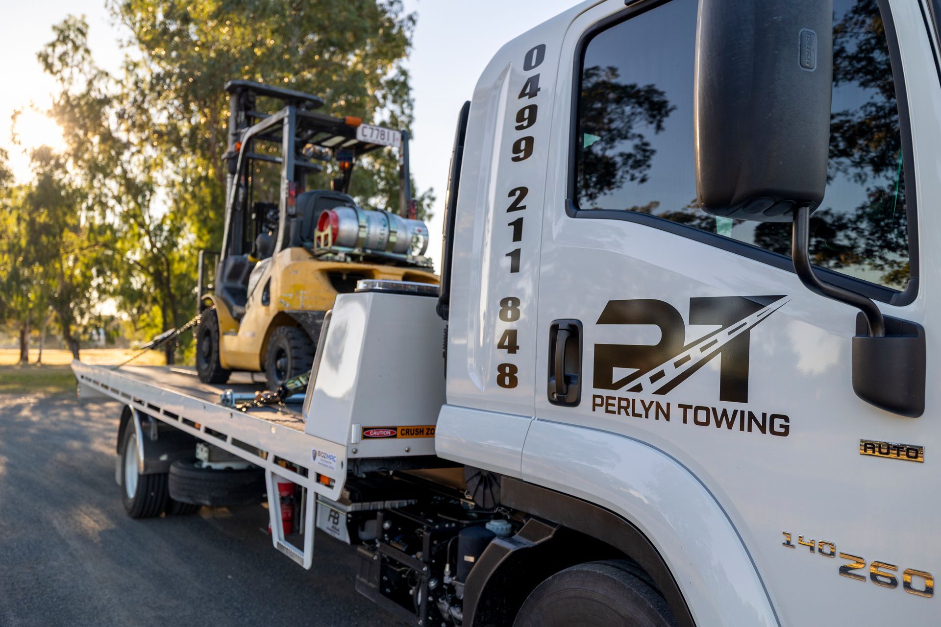 A white tow truck transporting a yellow forklift on its flatbed, with the sun setting in the background.— Perlyn Towing  in Rockhampton City, QLD