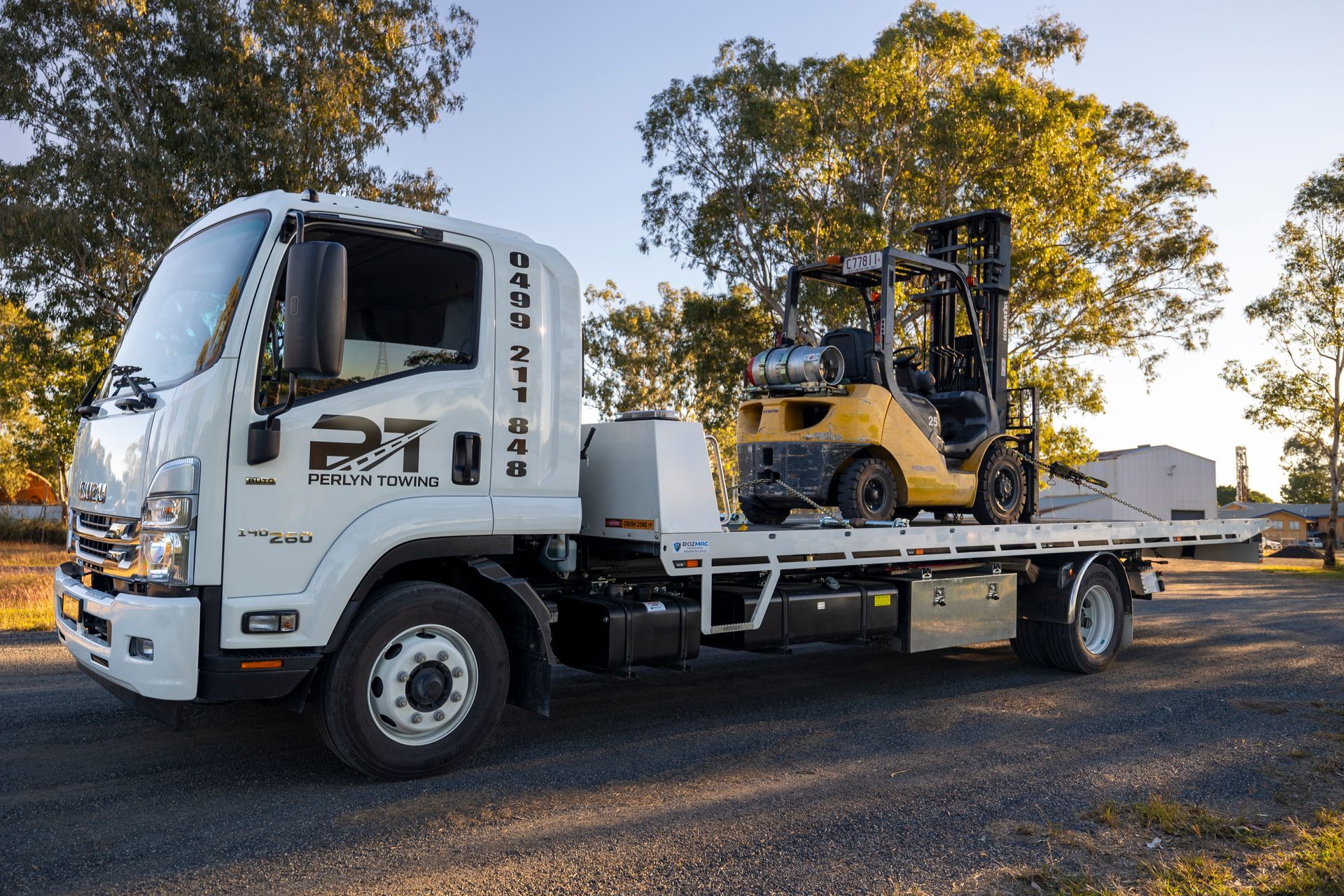 White truck carrying a yellow forklift on a flatbed, outdoors.— Perlyn Towing  in Gladstone, QLD
