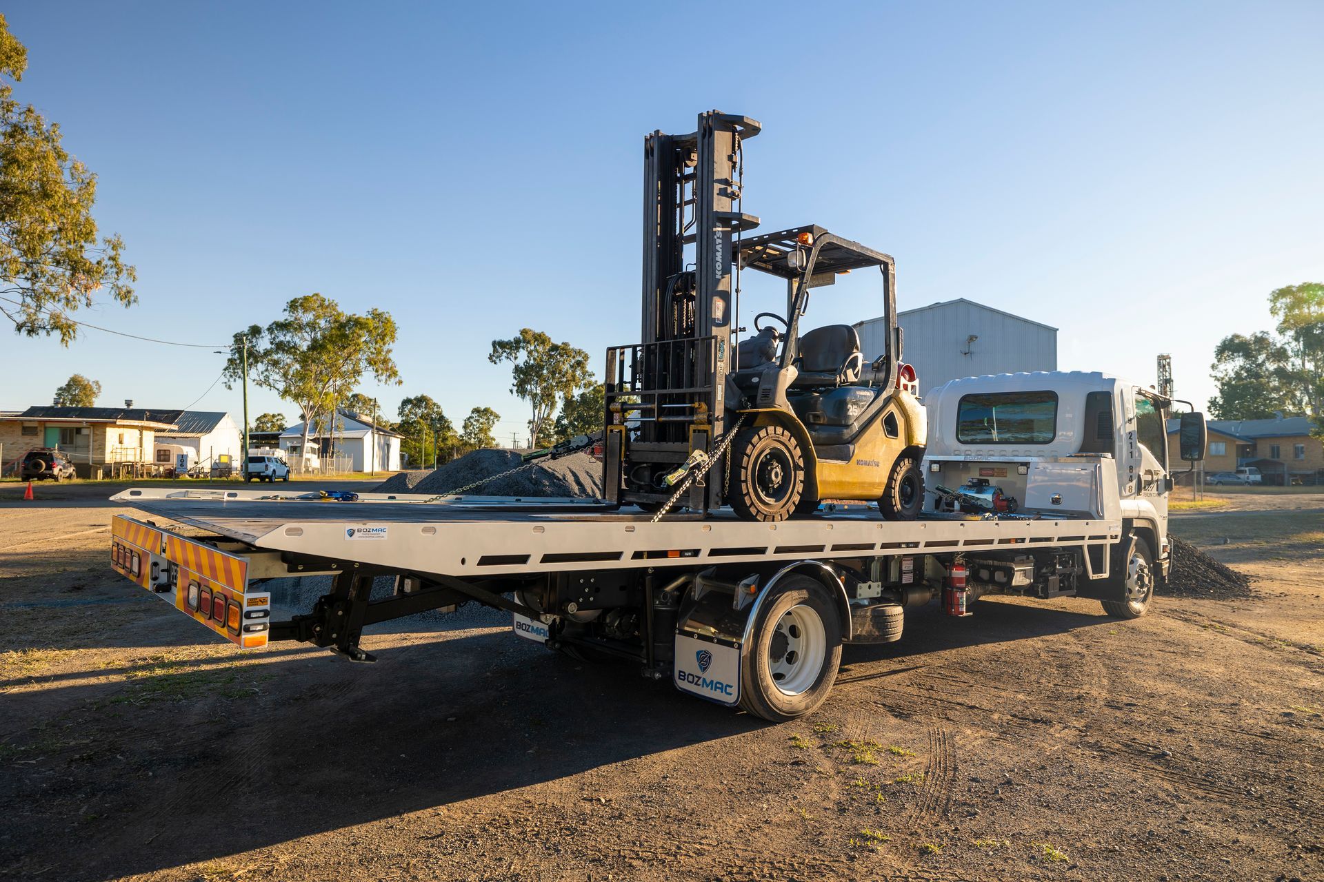 Yellow forklift loaded on a flatbed truck parked on dirt. Rural setting with houses and trees.— Perlyn Towing  in Gladstone, QLD