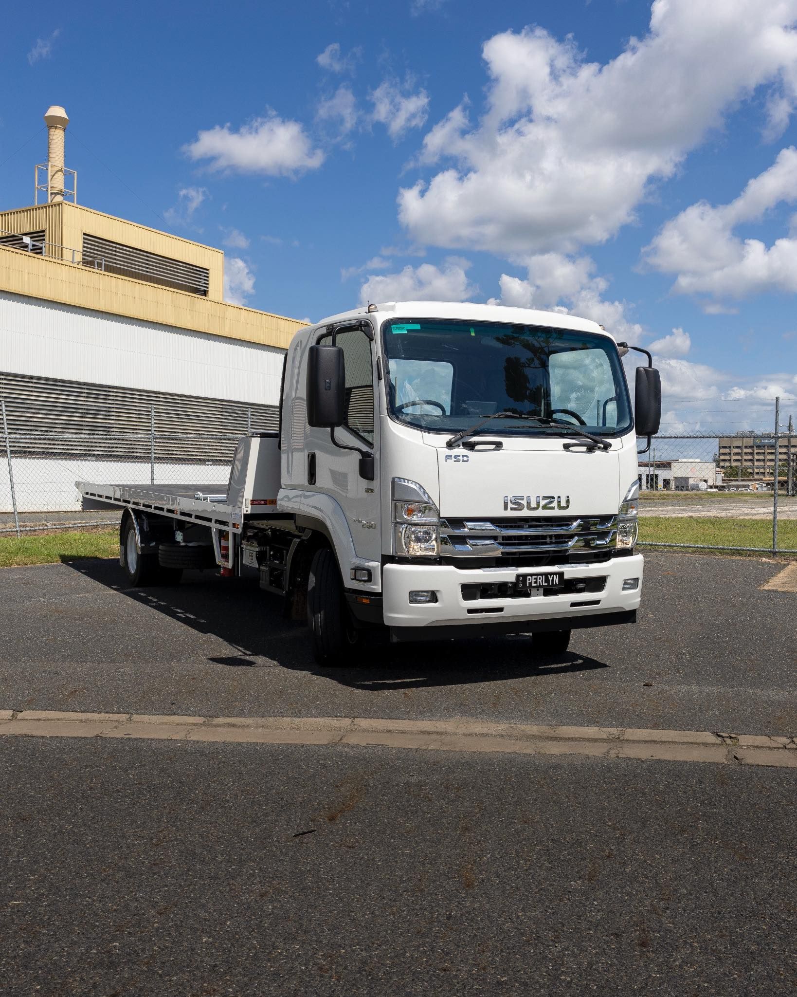 White Isuzu flatbed truck parked on asphalt in front of a building with a blue sky and clouds.— Perlyn Towing in Emerald, QLD