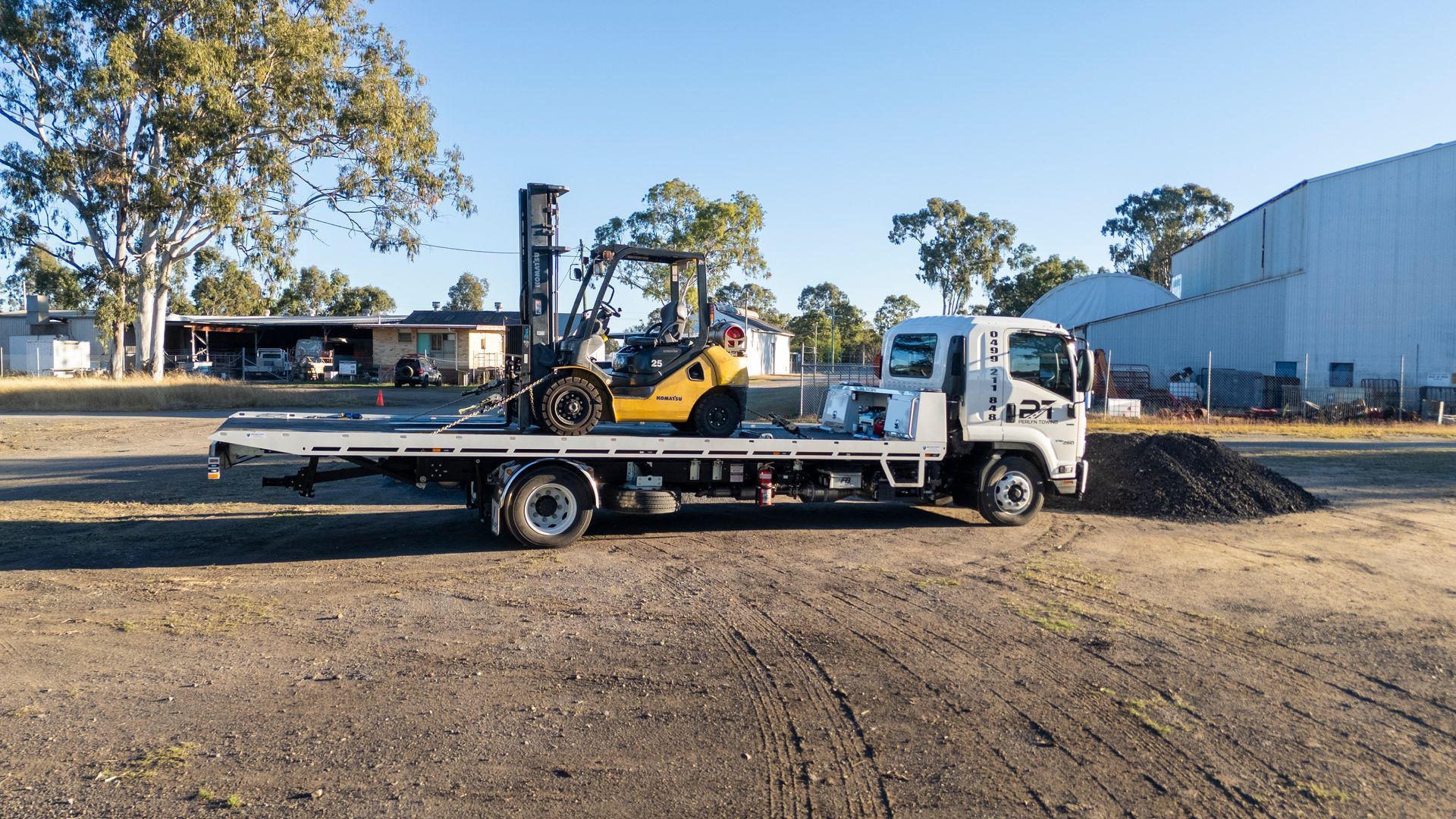 Flatbed truck carrying a yellow forklift on a gravel lot. Trees and a building are in the background.— Perlyn Towing  in Rockhampton City, QLD