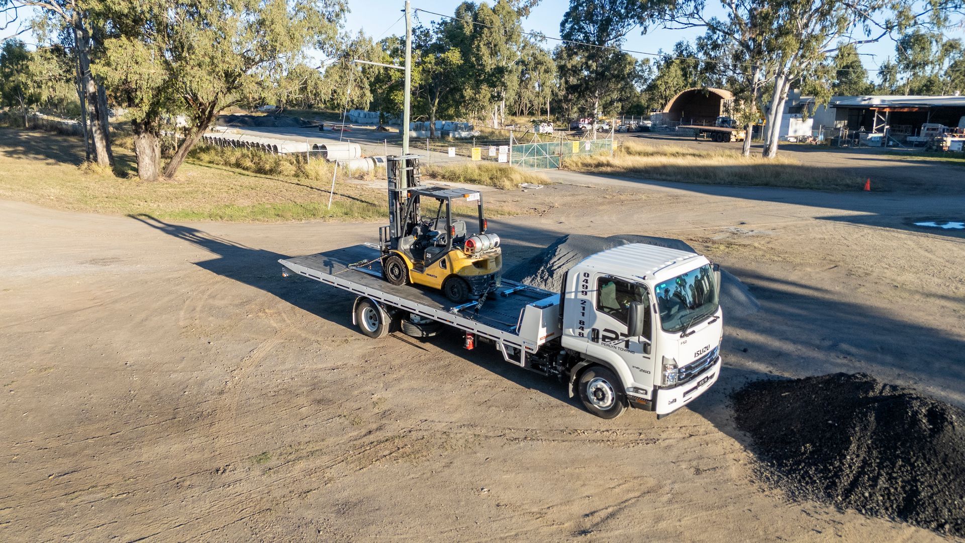 Flatbed truck carrying a yellow forklift in a rural area.— Perlyn Towing in Rockhampton City, QLD