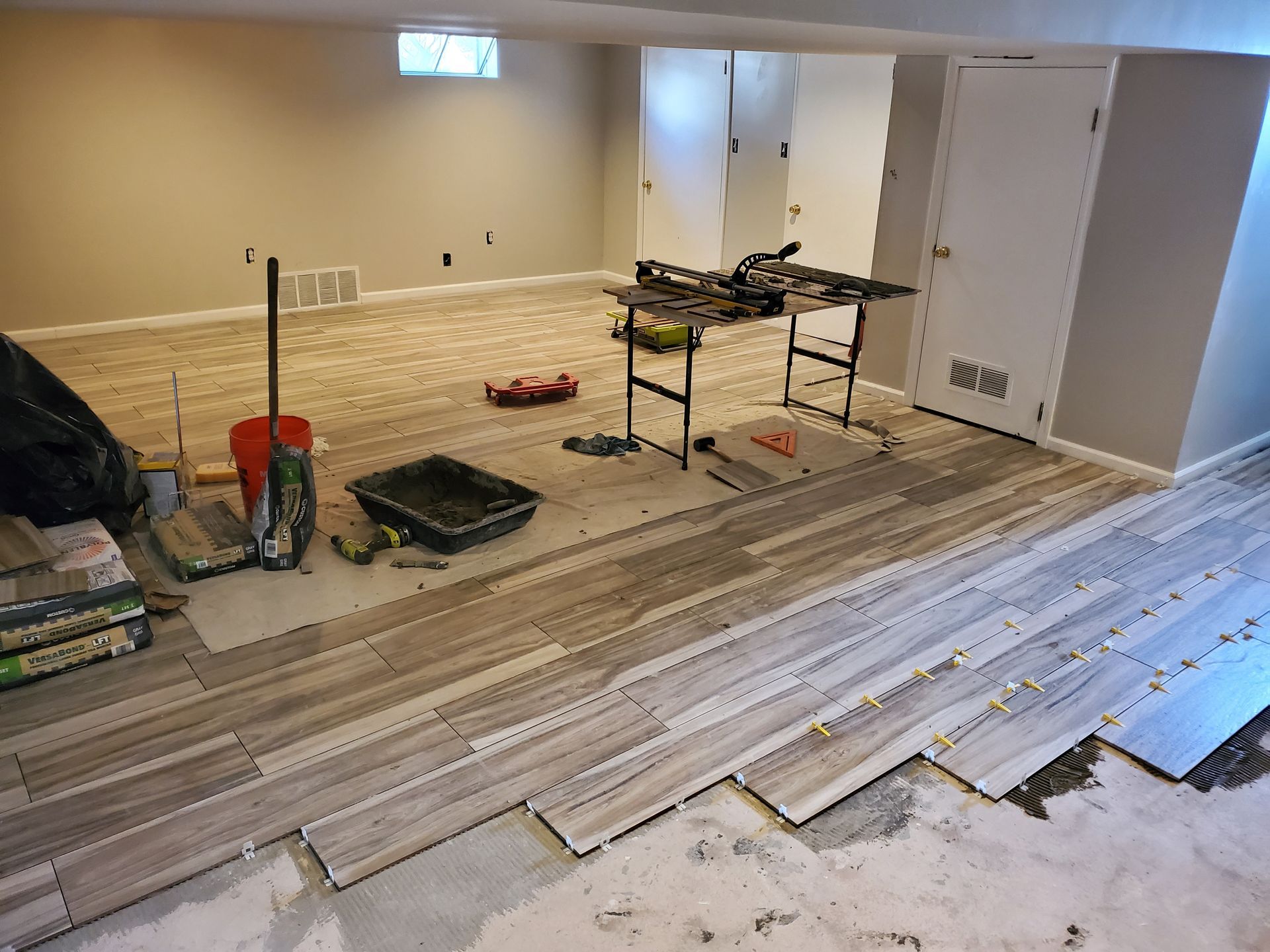Basement floor tiling in progress; worker's tools and partially laid wood-look tiles.