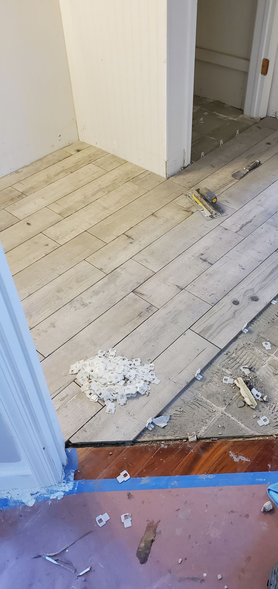 Tile flooring installation in progress; room interior with angled tile layout, spacers, and wood trim.